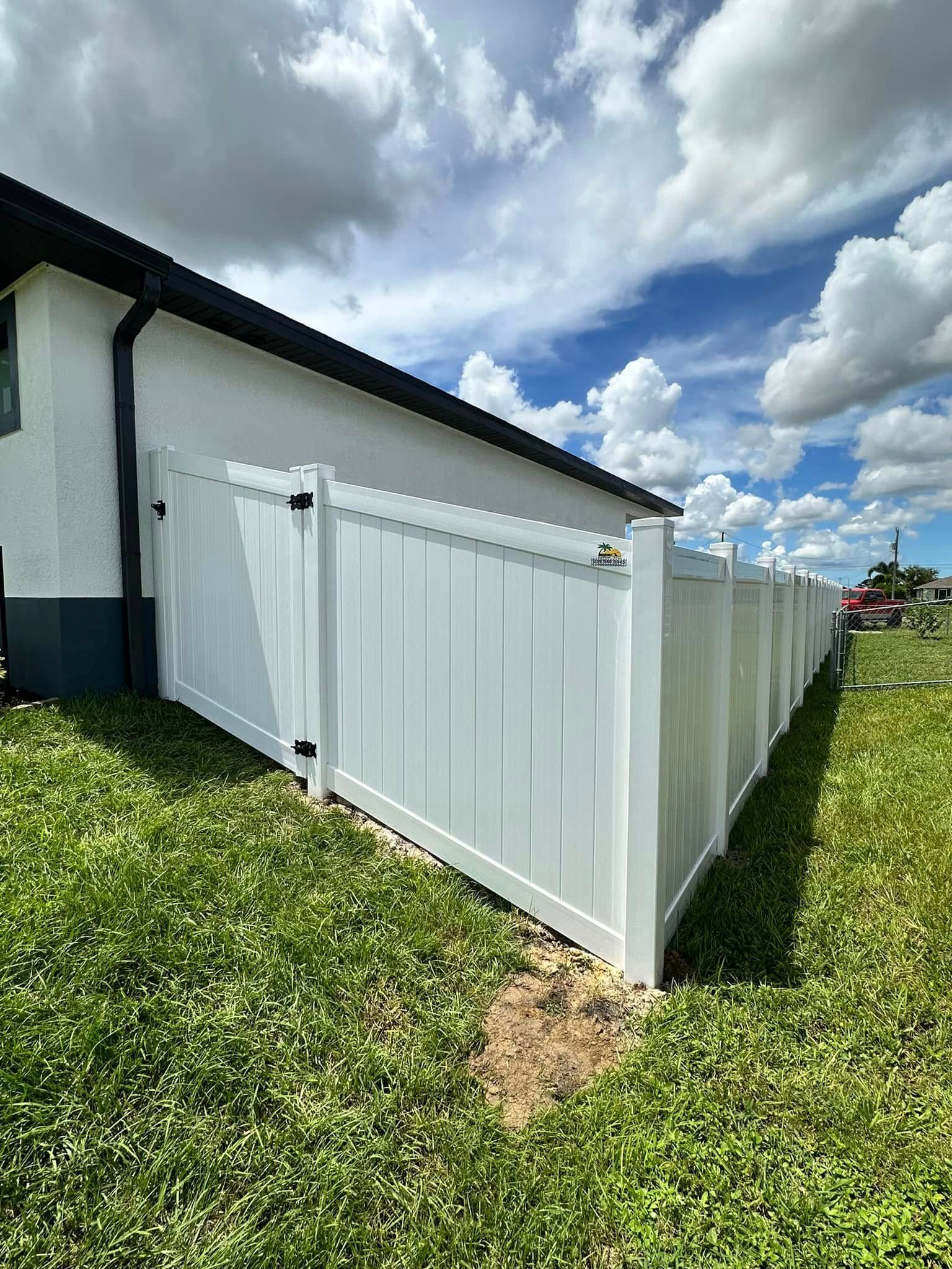 White vinyl fence bordering a green yard next to a white house under a cloudy blue sky.