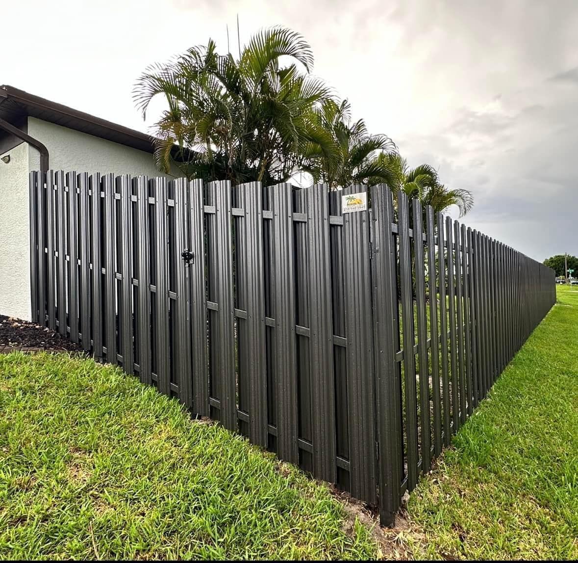 Black wooden privacy fence surrounding a green lawn, next to a beige house and palm trees.