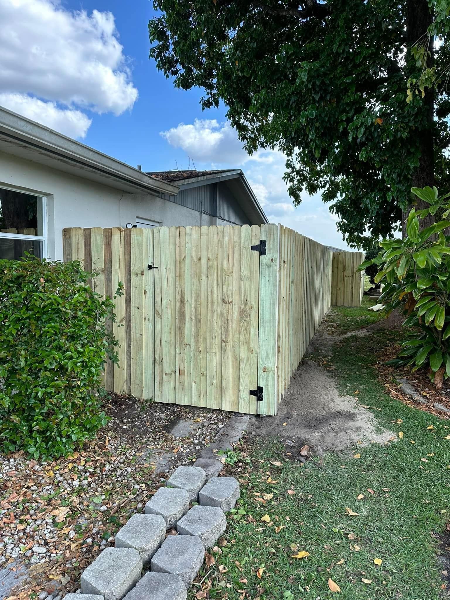 Wooden fence along a house and yard with greenery and a blue sky.