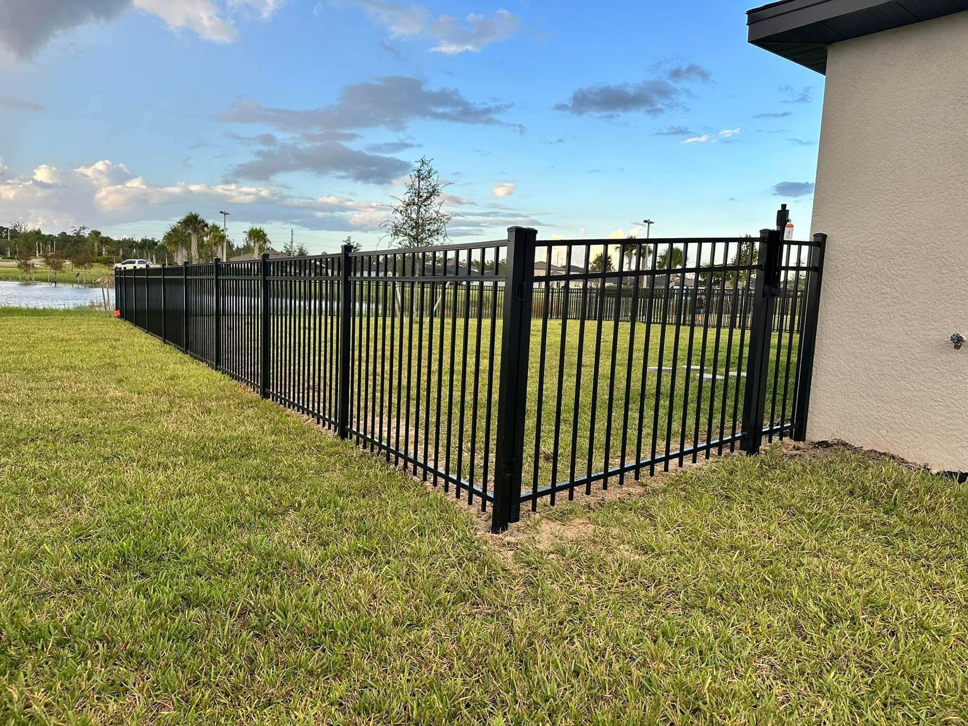 Black metal fence bordering a grassy yard near a lake and a house.
