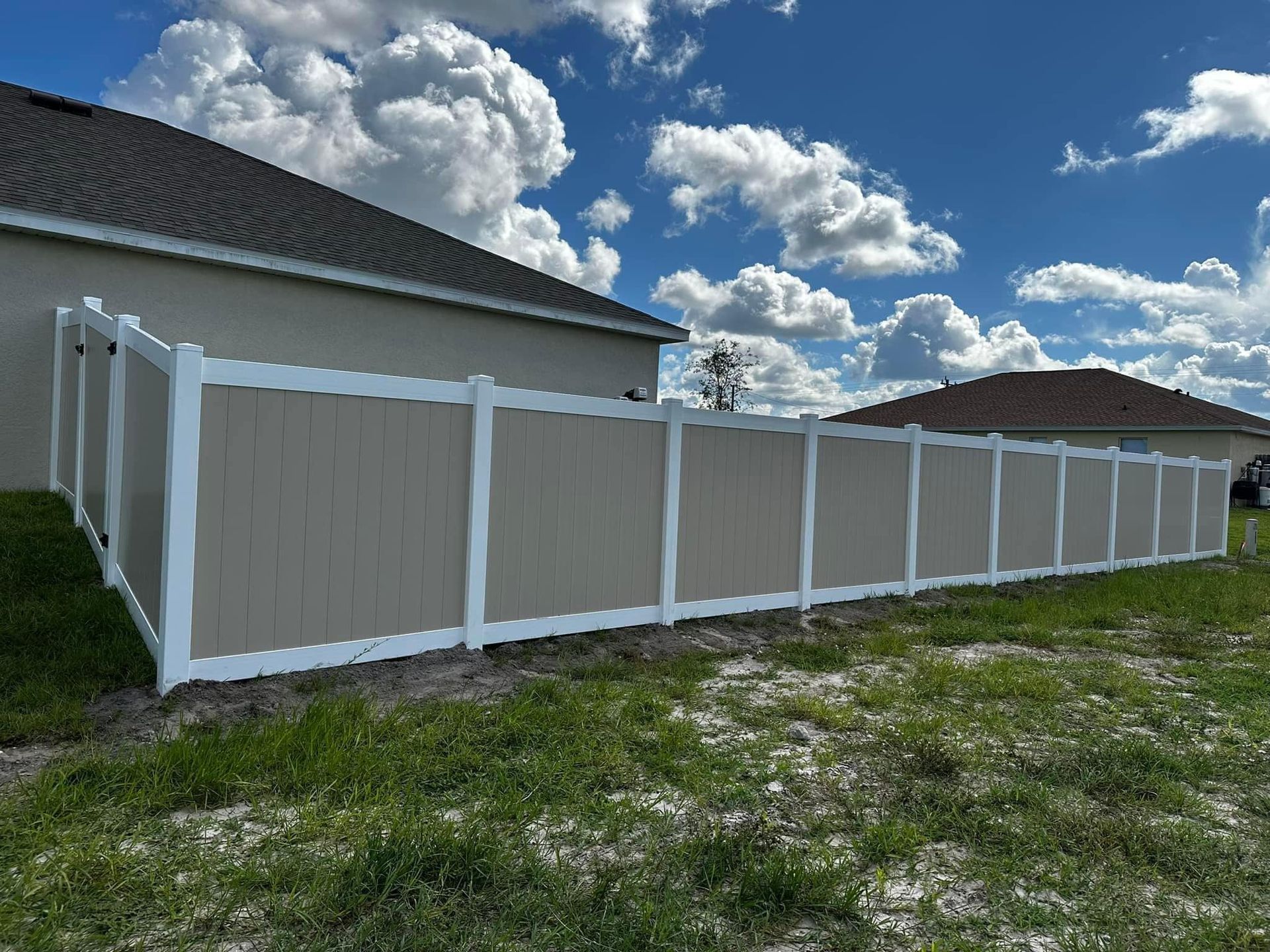 Tan and white vinyl fence between two houses under a partly cloudy blue sky.