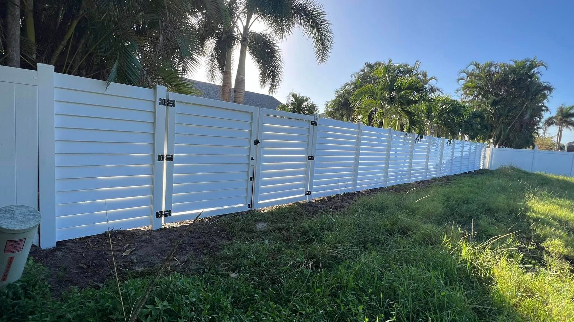 White horizontal slat fence in a yard with greenery and trees, under a bright blue sky.