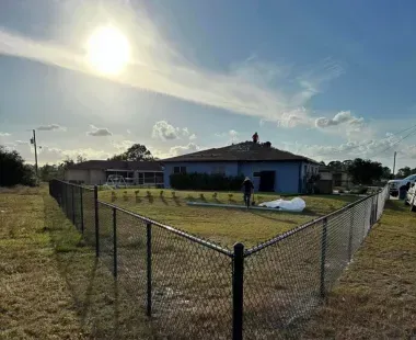 A chain link fence surrounds a grassy field in front of a house.