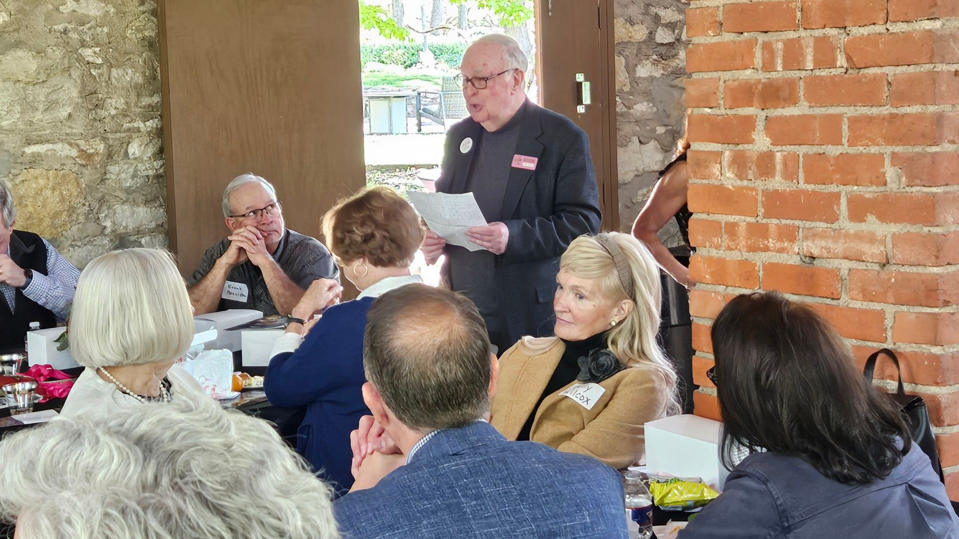A man reading from papers stands before a seated group at a table in a brick-walled room.