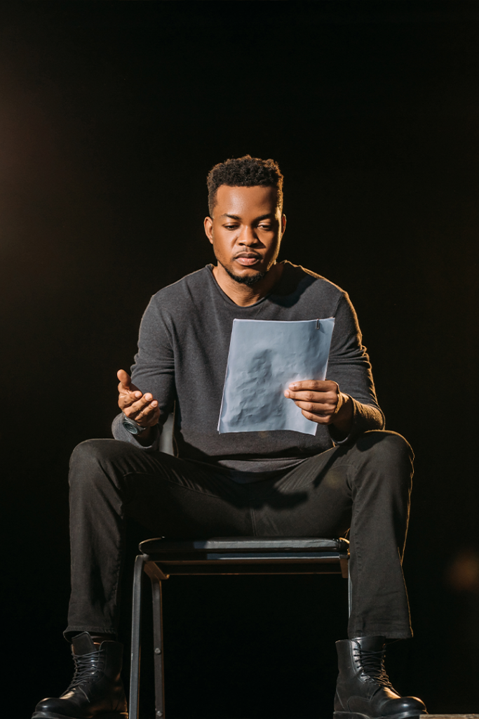 Man in a gray sweater, seated, holding a paper, looking down and gesturing. Black backdrop, dimly lit.