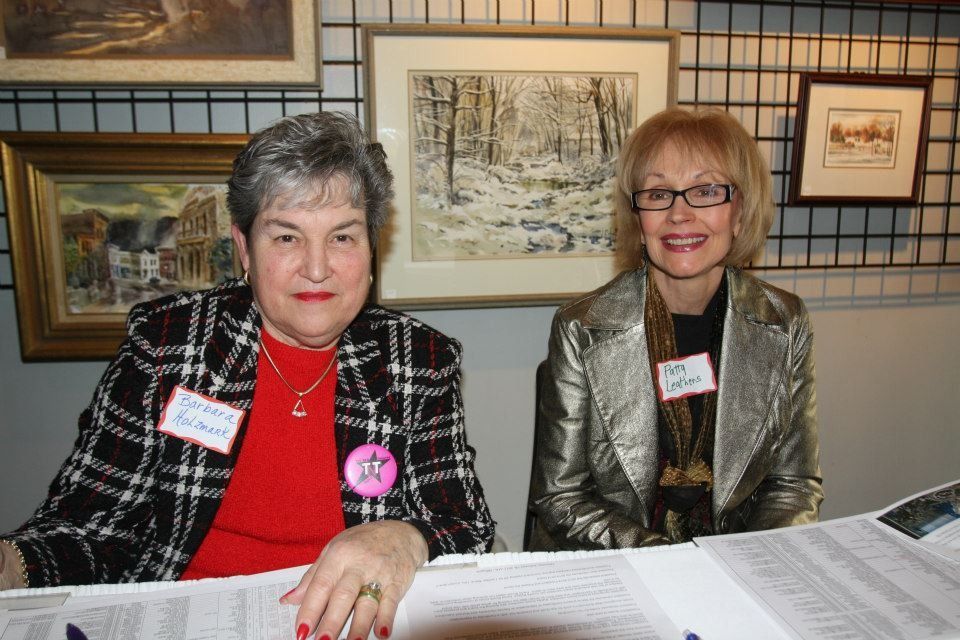 Two women behind a table with artwork in background. One in a red shirt with black and white jacket, other in a gold jacket.