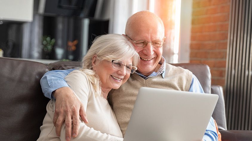 Smiling older couple sitting together on a couch, looking at a laptop
