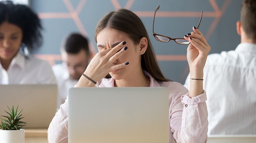 Woman in an office holding glasses and rubbing her eyes while sitting behind a laptop.