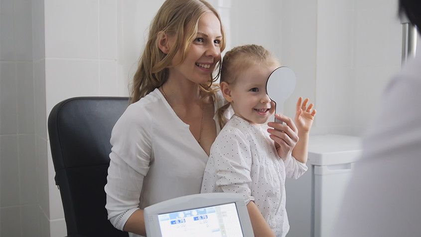Woman holding a smiling child in a medical office, with a doctor nearby and a tablet in the foreground