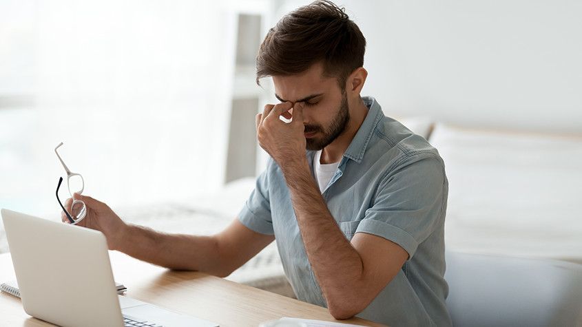 Man sitting at a desk with a laptop, rubbing his eyes while holding glasses, looking tired or stressed