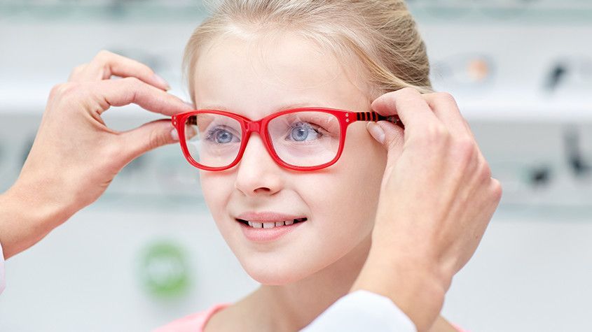 Person trying on bright red eyeglasses in a store, smiling as hands adjust the frames.