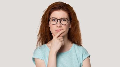 Thoughtful woman with glasses holding her chin, wearing a light blue shirt on a plain background