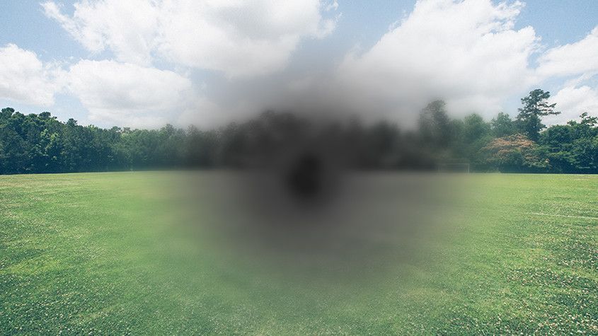 A grassy field with a blurry dark smoke plume rising near the center under a cloudy sky.