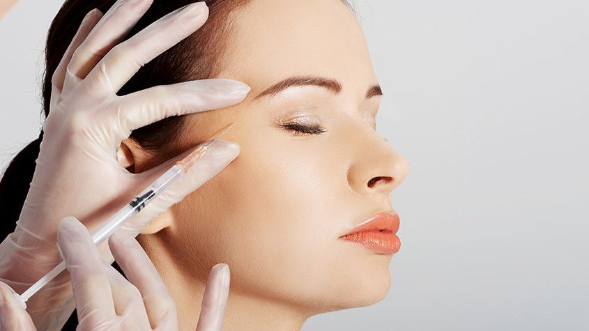 Cosmetic procedure in progress on a woman’s face with gloved hands and a syringe near her cheek