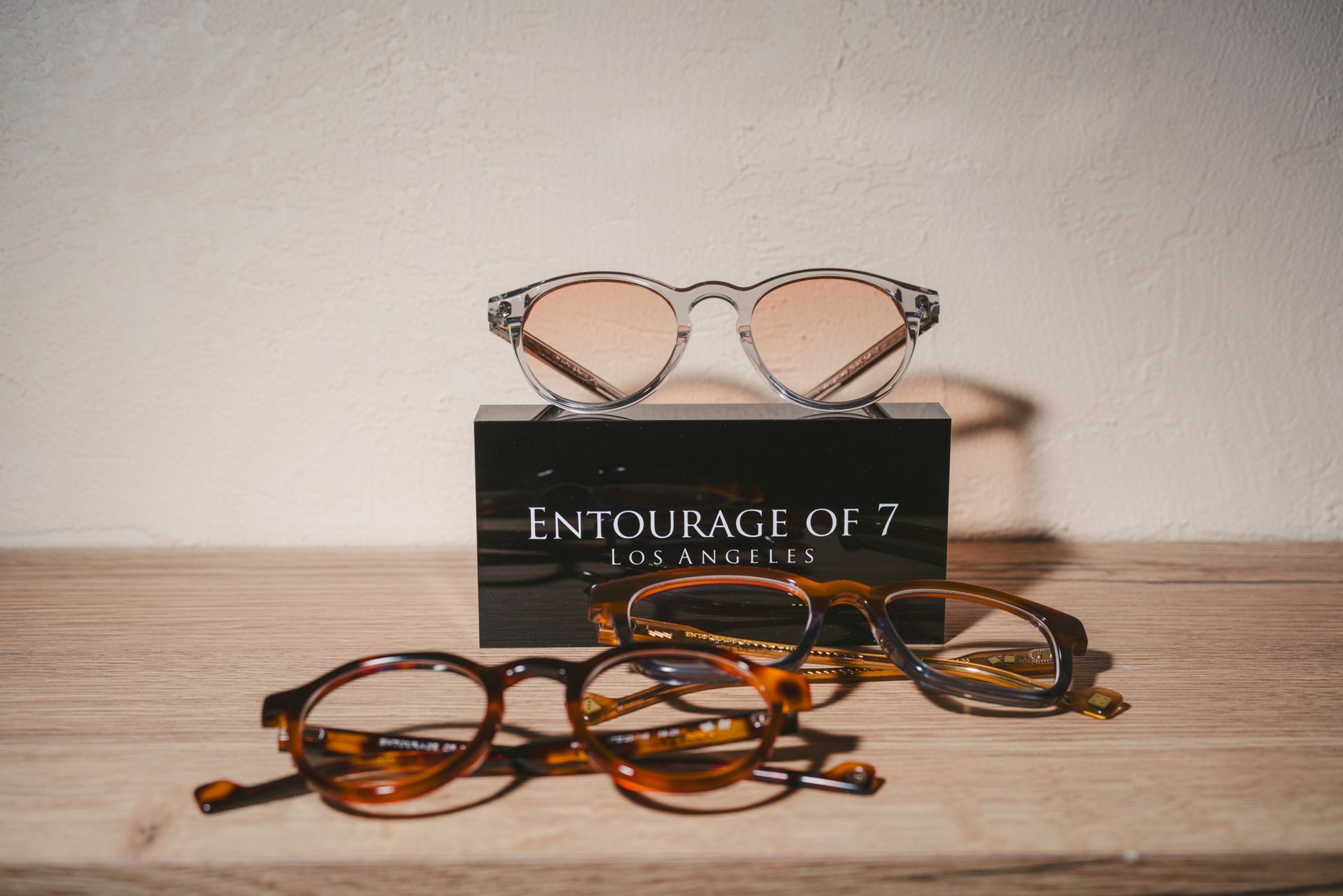 Three pairs of eyeglasses stacked on a book titled “ENTOURAGE OF 7” on a wooden table.