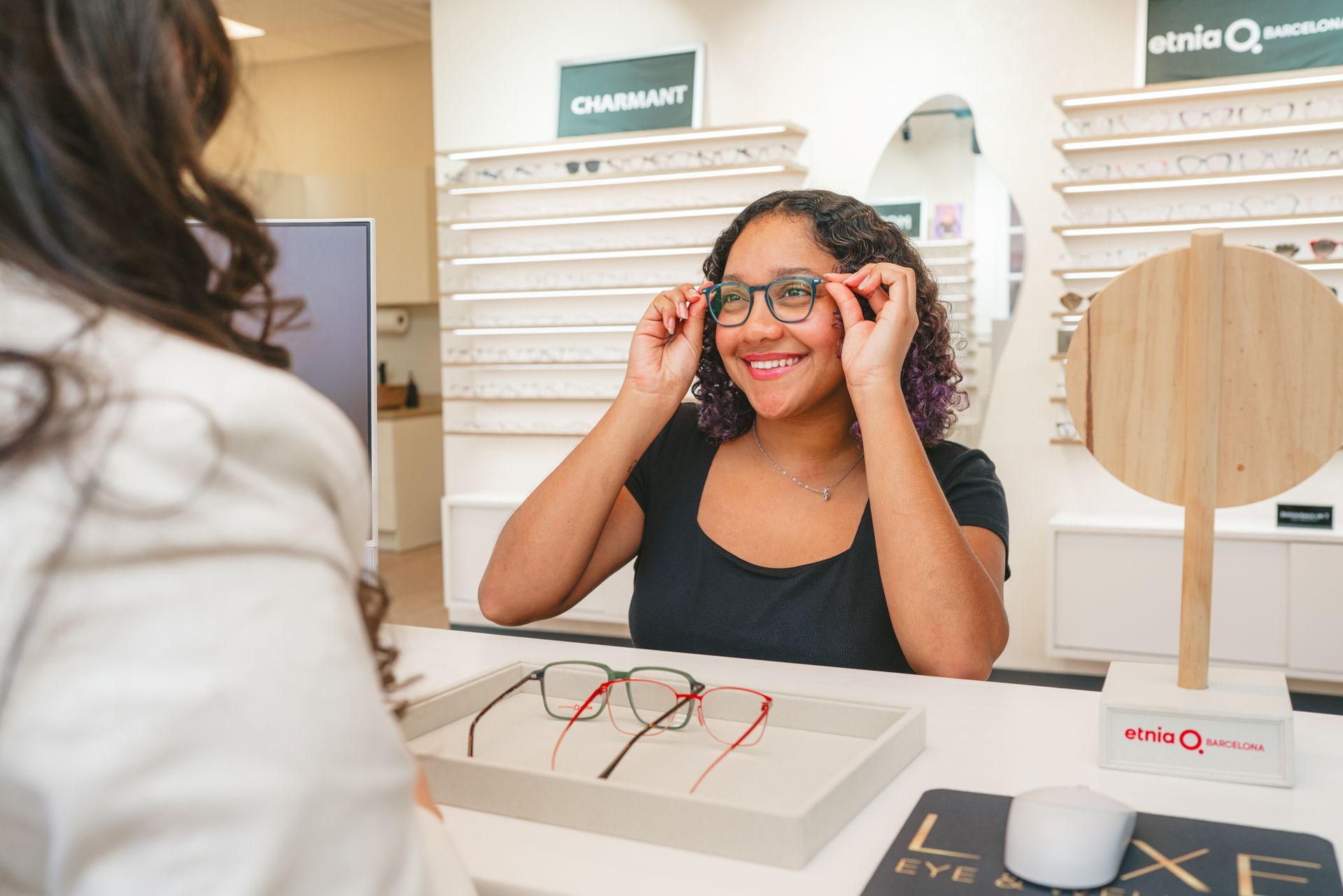 Woman trying on glasses at an eyeglasses store counter, smiling at her reflection