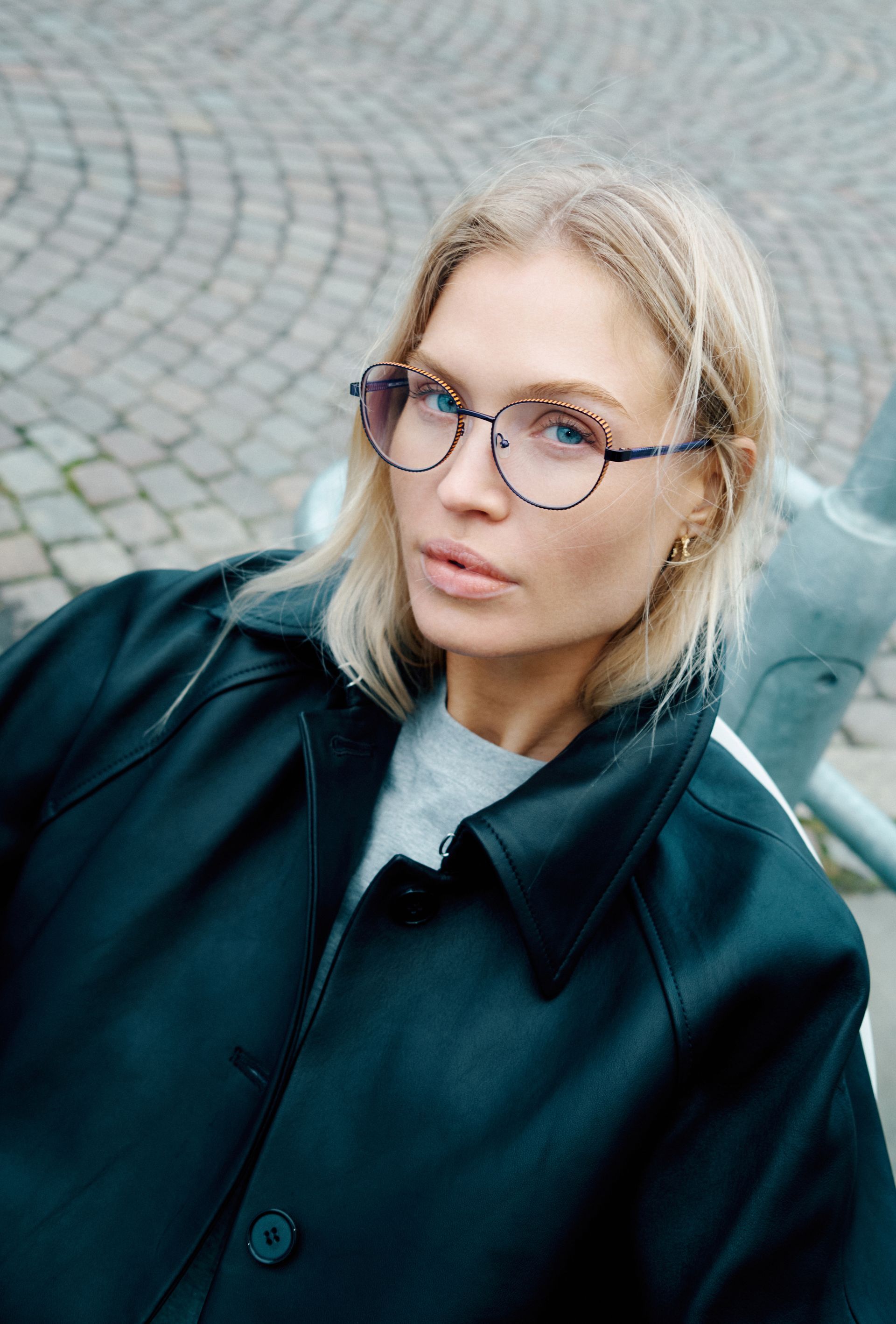 Person with blond hair and glasses wearing a dark coat, looking at the camera on a cobblestone street.