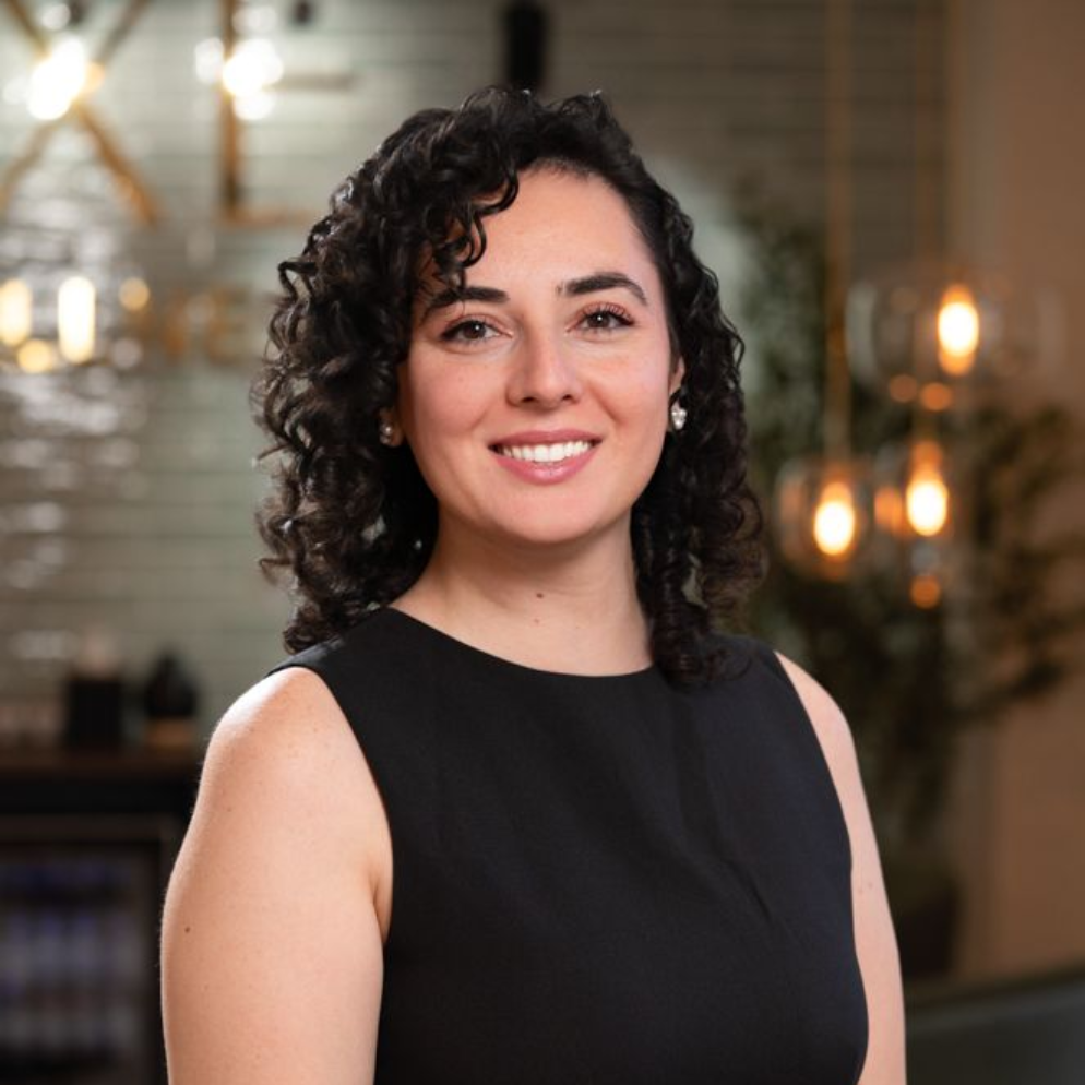 Portrait of a smiling person with curly dark hair, wearing a black top, against a softly blurred indoor background
