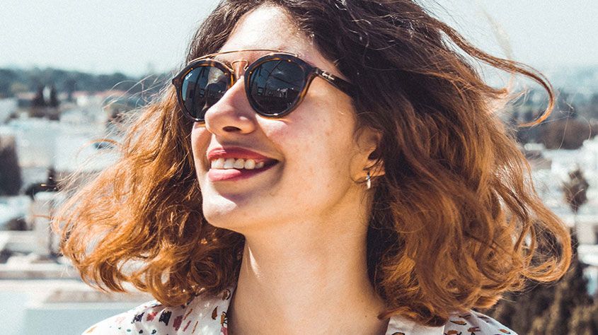 Smiling woman wearing sunglasses, with wavy hair, outdoors in bright sunlight.
