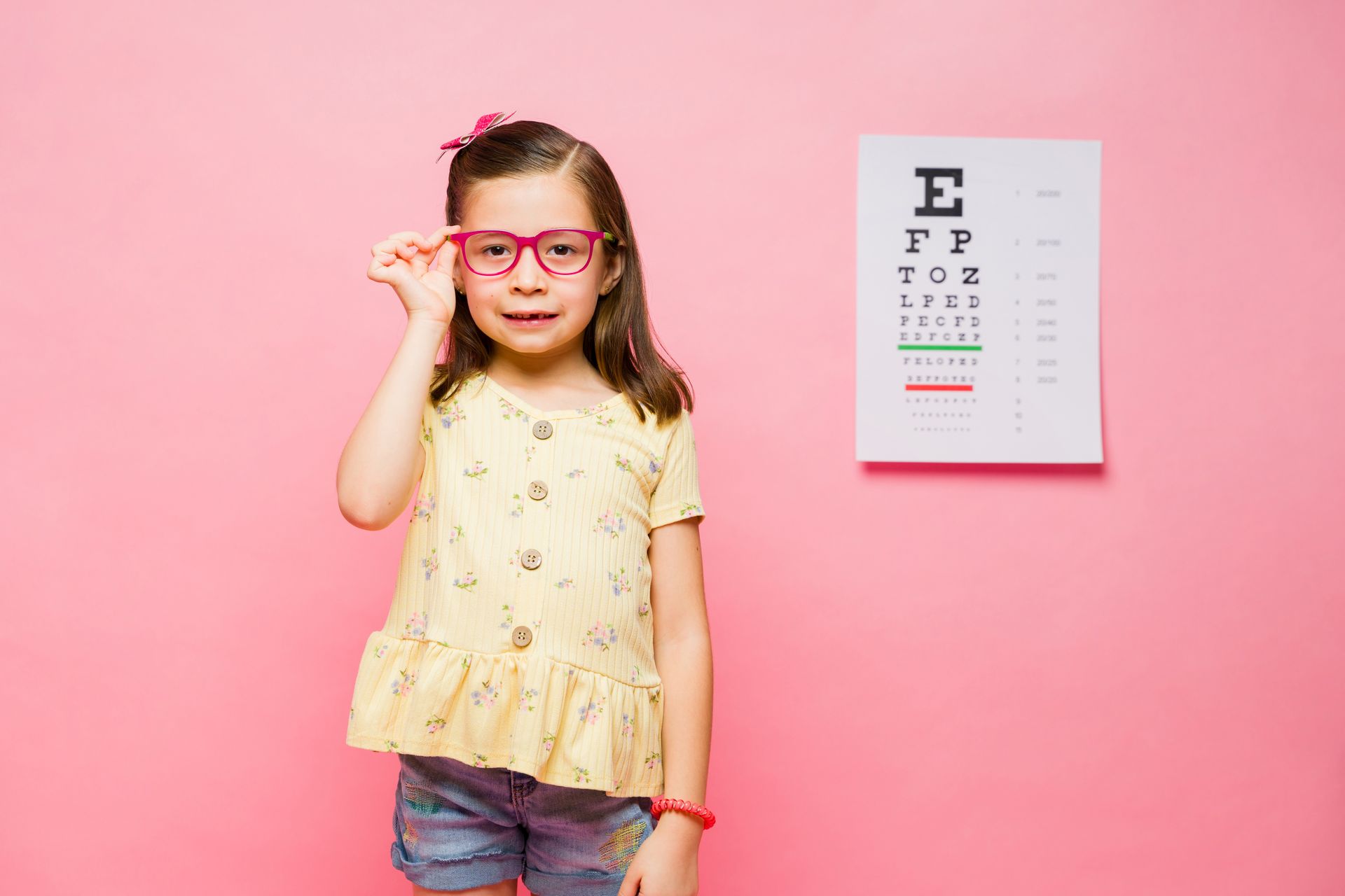 Child in pink glasses adjusts frames beside a vision chart on a pink wall