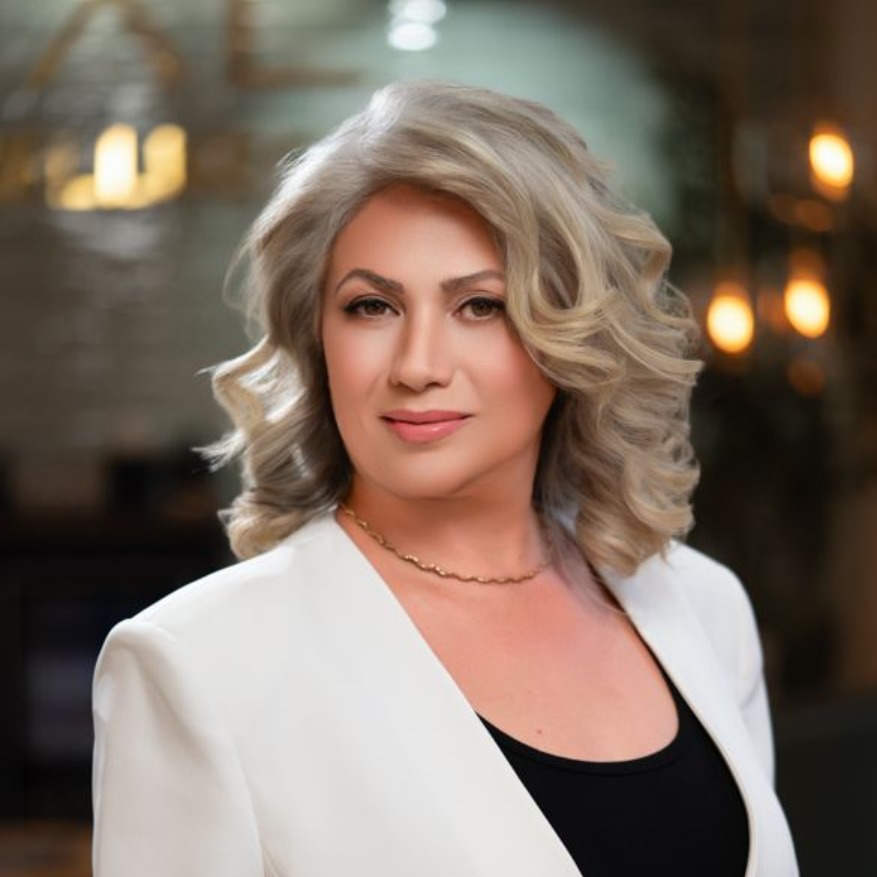 Portrait of a woman with wavy blonde hair in a white top, softly lit against a blurred background