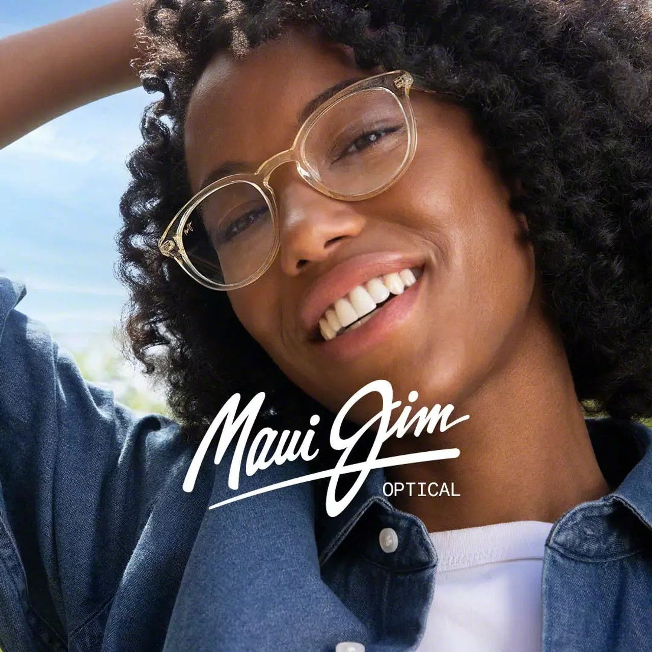 Smiling person wearing gold glasses in a Maui Jim Optical ad, with arm raised against a bright sky