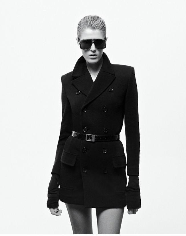 Model in black coat and sunglasses, standing against a white background.