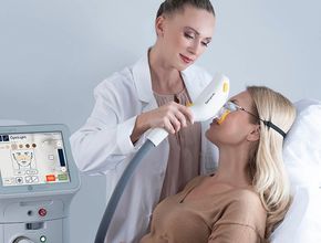 A clinician uses a handheld device on a seated patient beside a facial treatment machine.