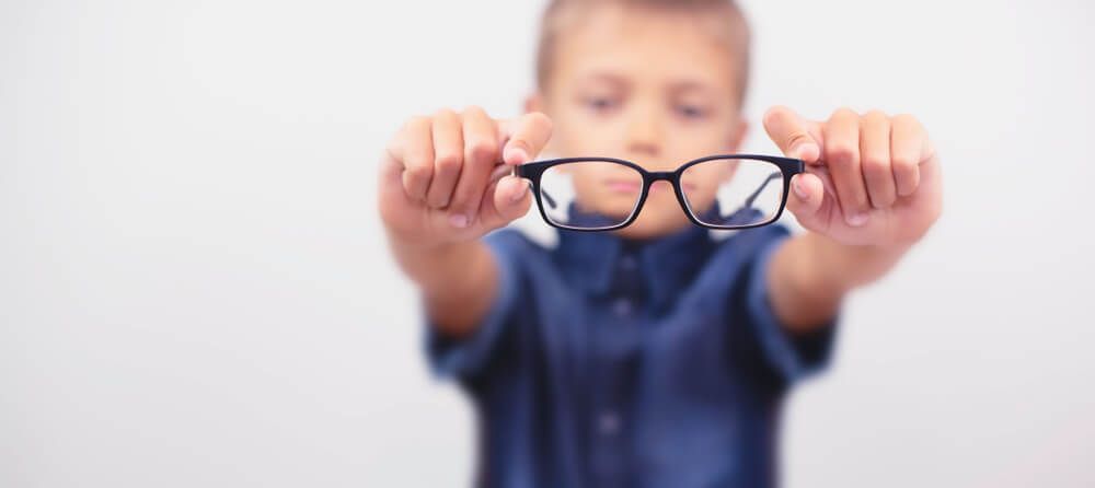 Child holding black eyeglasses toward the camera against a blurred light background