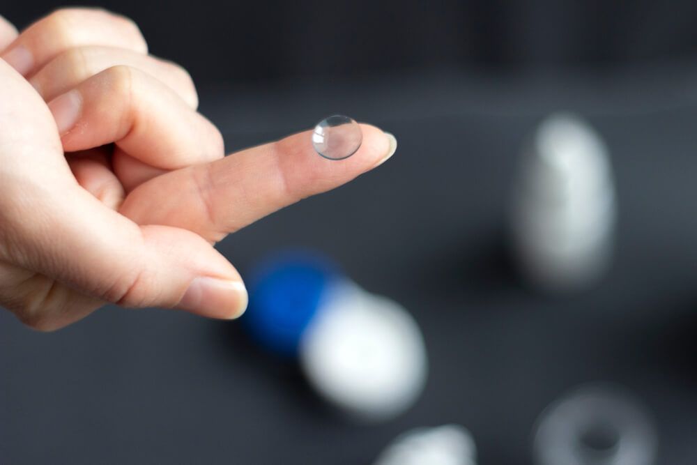 Hand holding a clear contact lens with blurred contact lens cases in the background