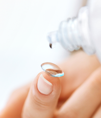 A contact lens held on a fingertip with a drop from a lens solution bottle above it