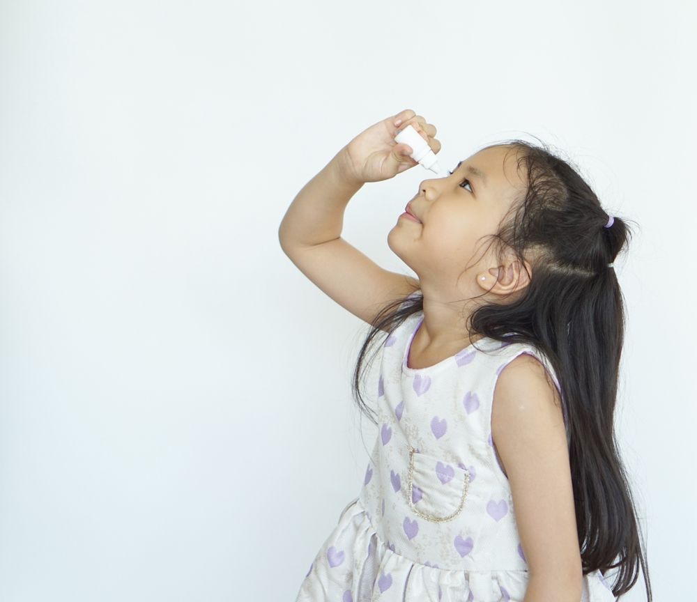 Young child in a white dress looking up and holding a small object near her face against a plain background