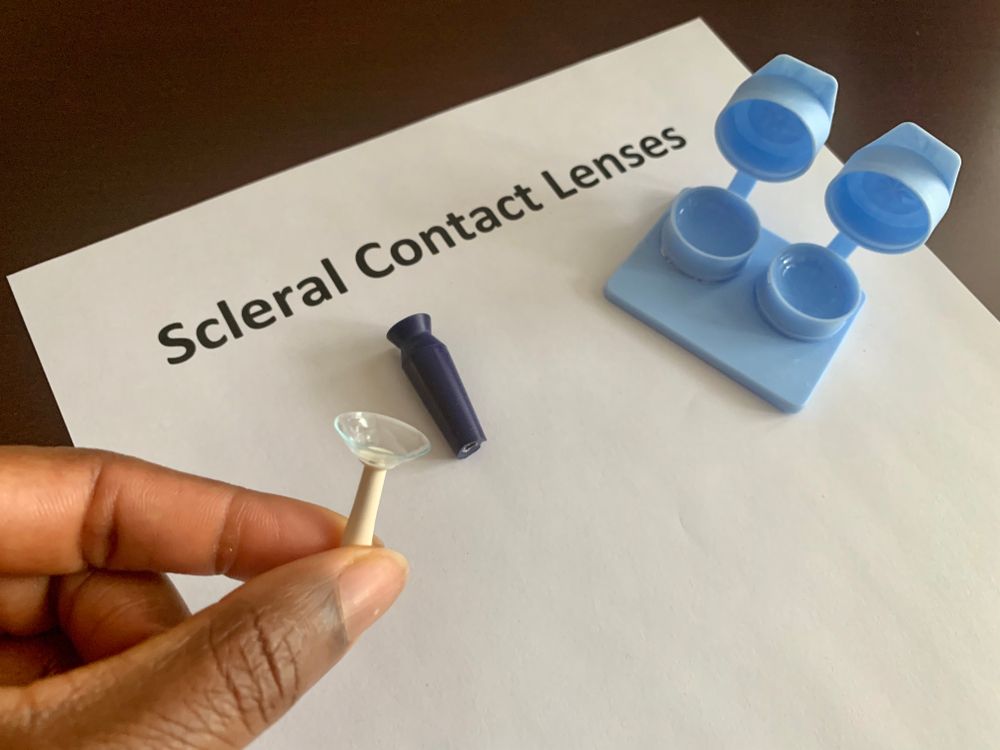Hand holding a clear scleral contact lens and applicator beside labeled lens case and tools