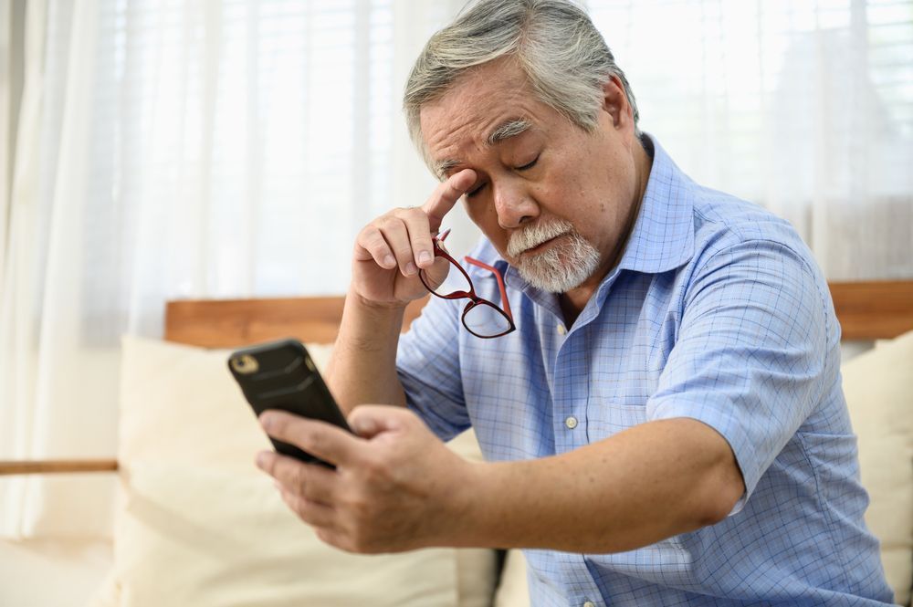 Man rubbing his eyes while looking at a smartphone on a couch in a bright room