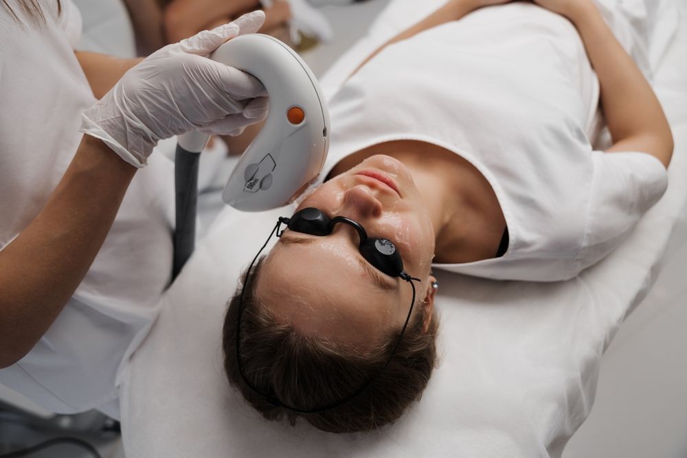 Person wearing protective glasses reclines on a treatment bed while holding a handheld facial device.
