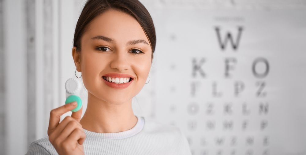 Person holding a contact lens beside an eye chart, smiling in a bright clinic setting