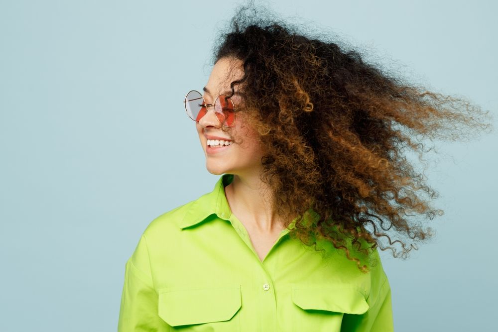 Smiling person in bright green shirt with curly hair blowing against a light blue background