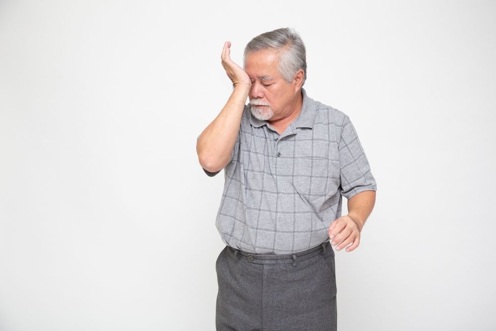 Older man touching his face with one hand, standing against a plain white background.