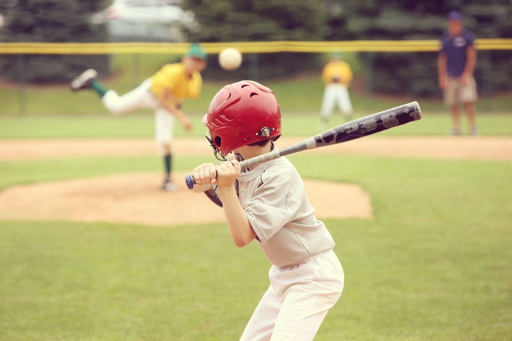 Young baseball player in red helmet batting on a field, with pitcher blurred in the background