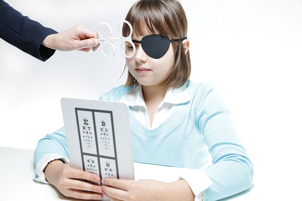 Person wearing an eye patch reads an eye chart while another hand holds up an eye test lens.
