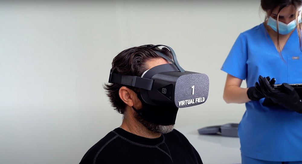 Person wearing a black VR headset while a clinician in blue scrubs stands nearby.