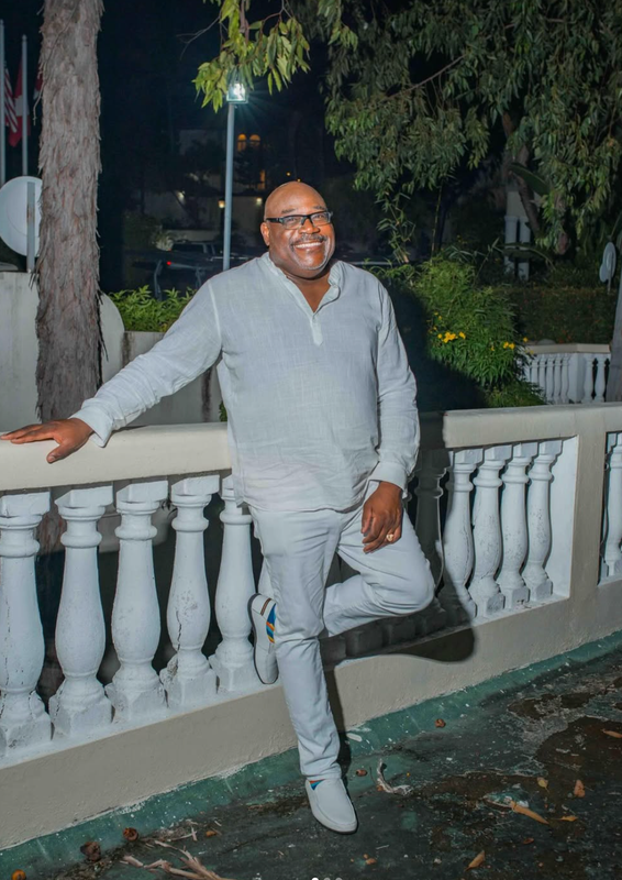 Man smiling, leaning on a white railing at night outdoors, wearing a light gray outfit.