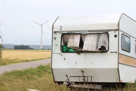 Old, damaged white camper trailer in a field with a road, yellow field, and windmills in the background.