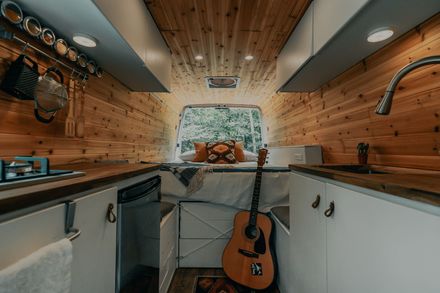 Cozy camper van interior with wood paneling, bed, guitar, and kitchen area.