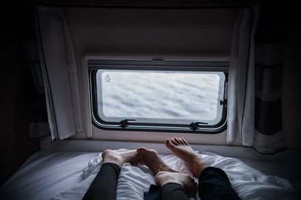 Two pairs of feet on a bed in front of a window, looking out at water.