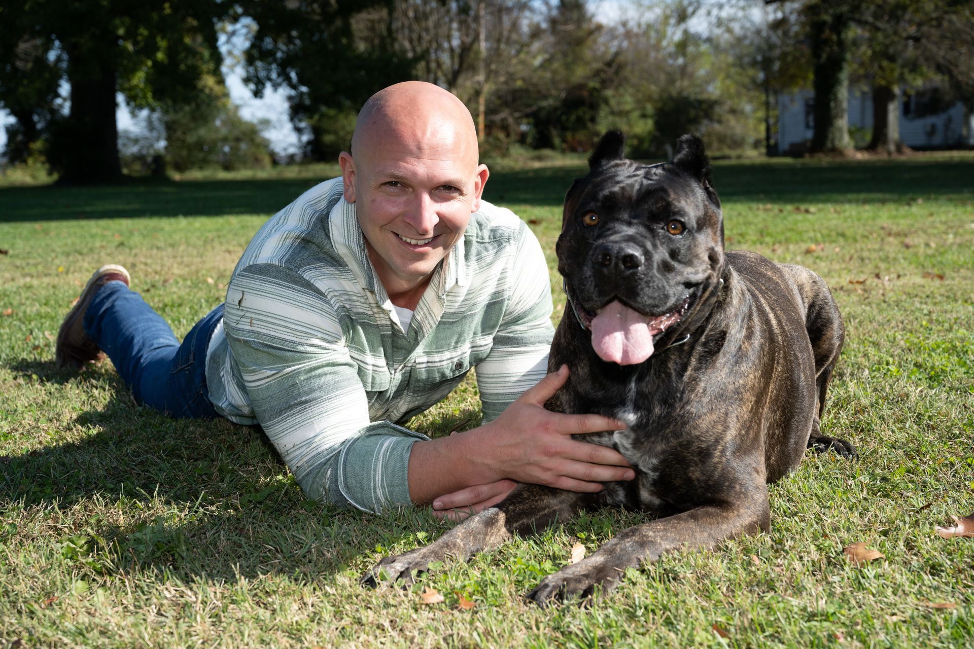 Man lying on grass beside a black dog with tongue out in a sunny park