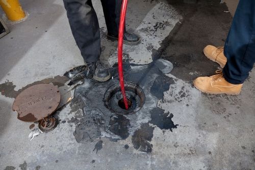 Workers unclogging a floor drain with a red hose amid water and debris on a concrete floor