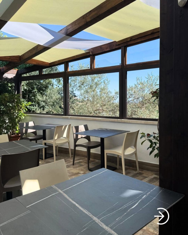 Terrazza soleggiata all'aperto con tavoli e sedie sotto una tettoia in vetro e legno, con vista sugli alberi.