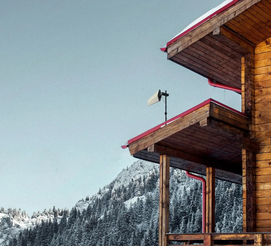 Wooden alpine cabin with red-trimmed roof overlooking snowy pine-covered mountains under a clear sky