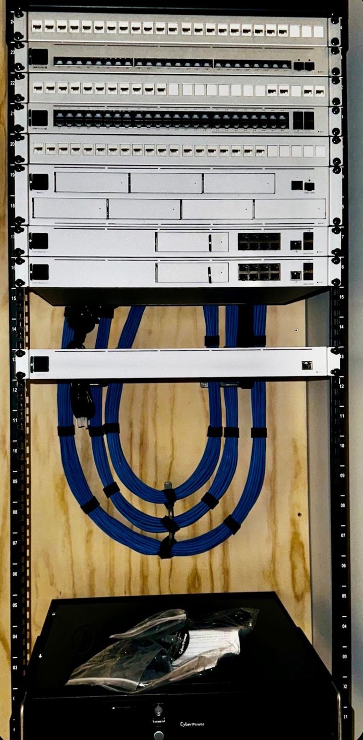 Wall-mounted network rack with patch panels and blue Ethernet cables above a black equipment box